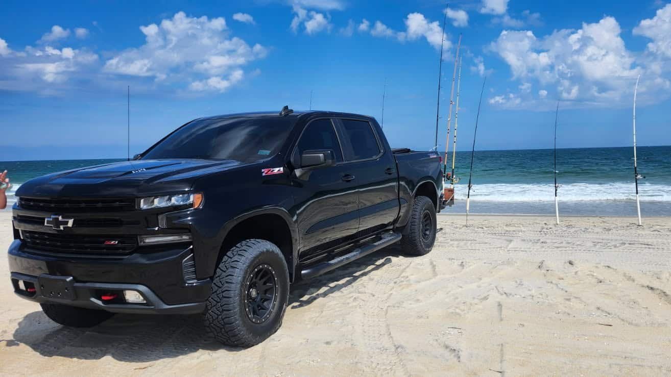 Real photo: Beach 4x4's black Chevrolet Silverado Trail Boss parked on the beach with surf-fishing rods in the sand.