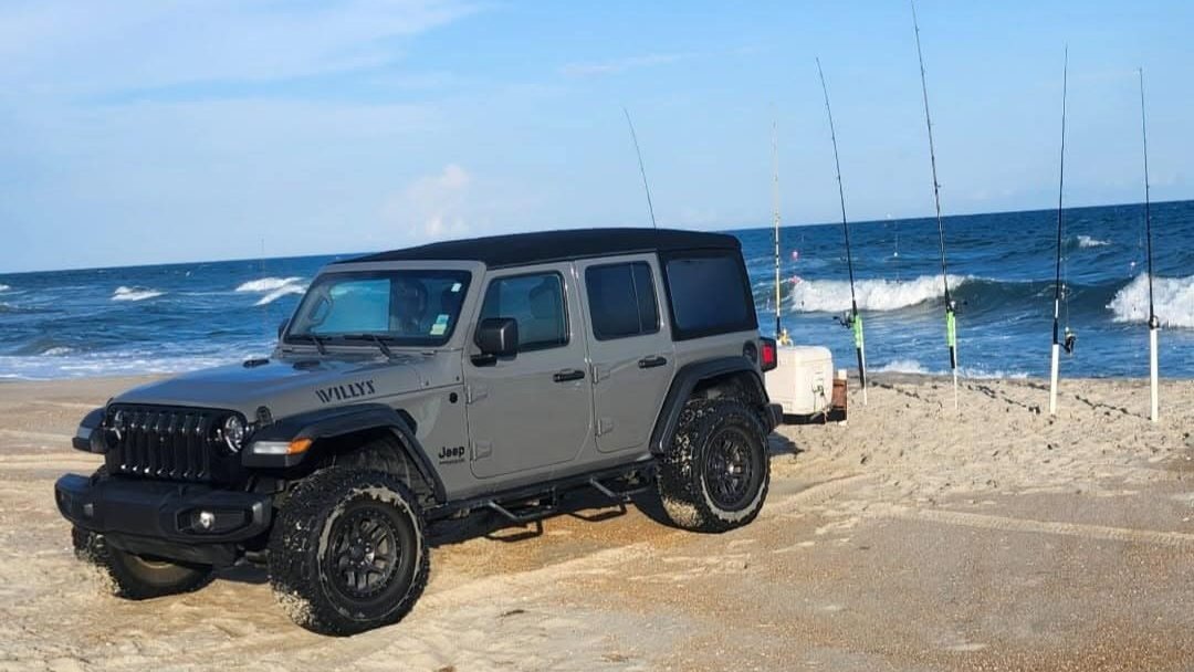 Real photo: a gray Beach 4x4 Jeep Wrangler Willys on the beach with surf-fishing rods in the sand.